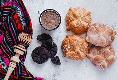 Cuatro panes de muerto junto con una taza de chocolate, todos cocinados con cardamomo